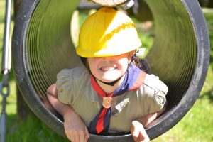 Cub with necker in a tube on assault course with hard hat covering her eyes